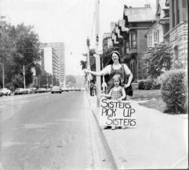 Woman and little girl standing on sidewalk and holding a "Sisters pick up sisters" sign
